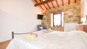 Un dormitorio con pared de piedra, una cama con ropa de cama blanca, un sillón gris, una ventana con cortinas y un televisor montado en la pared.
