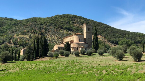 Un edificio histórico con un campanario, rodeado de olivares y cipreses en una colina.