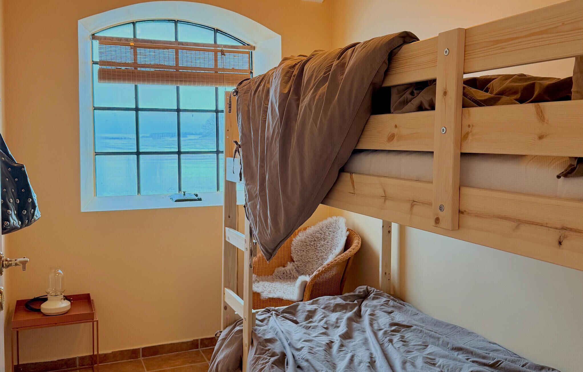 Una cama individual con cabecera de madera, una silla de mimbre, una mesita con lámpara y una ventana con persianas.