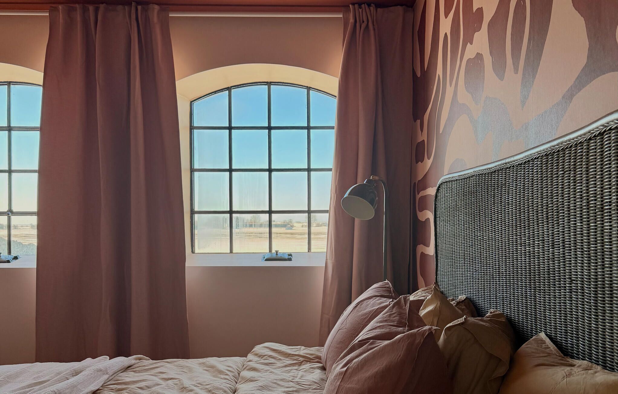 A bedroom with a large window, a bed with a headboard, and a lamp.