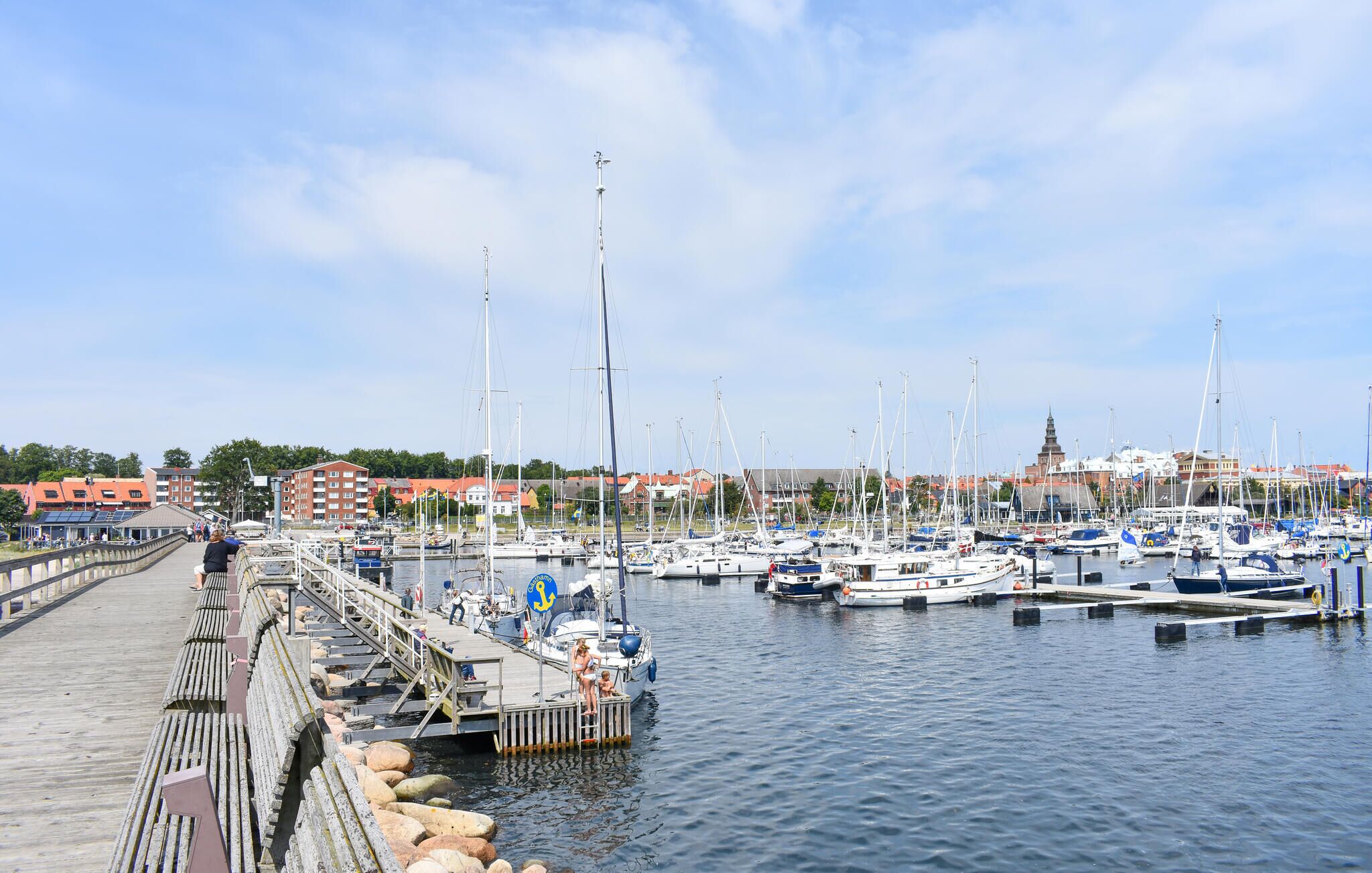 A marina with boats docked, a pier with people, and buildings in the background.
