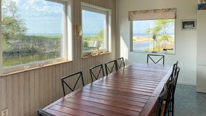 Un comedor con una mesa y sillas de madera, ventanas amplias con vistas a la playa y al agua, y un estante pequeño con una lámpara.