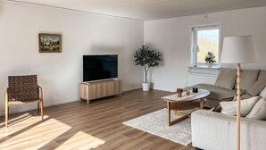 Ein Wohnzimmer mit Fernseher, einer Couch, einem Couchtisch, einem Sessel, einer Pflanze und einem Fenster.