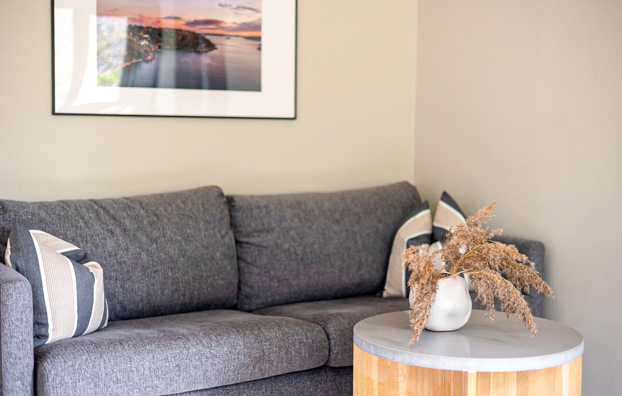 A grey sofa with striped pillows, a round wooden side table with a white vase and dried plants, and a framed picture on the wall.