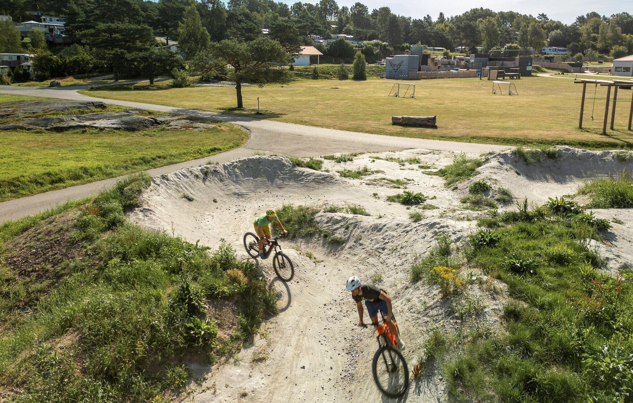 A dirt bike track with two riders, surrounded by greenery and houses in the background.