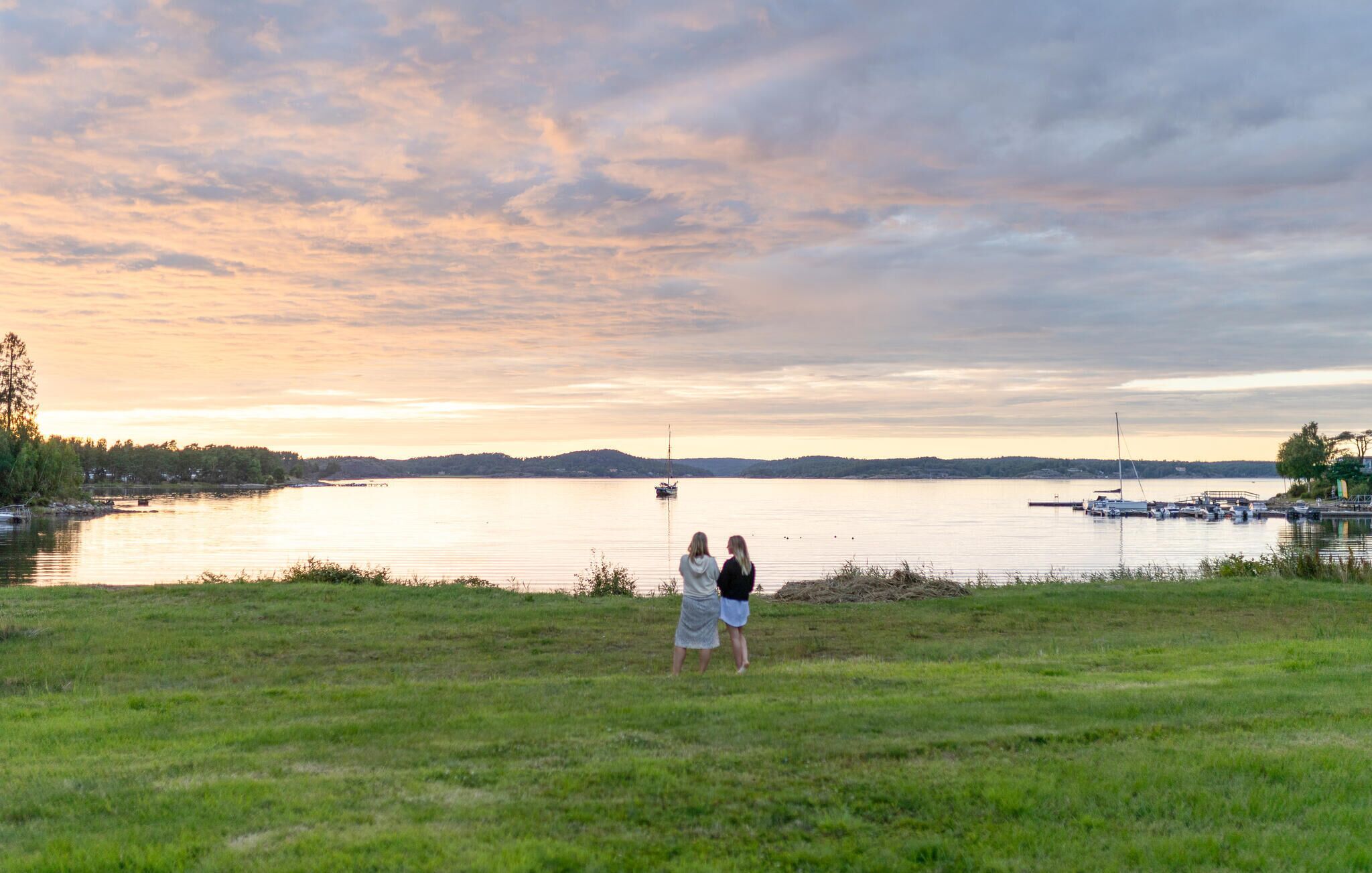Un campo cubierto de césped con dos personas de pie, contemplando un lago tranquilo con barcos y una orilla distante.