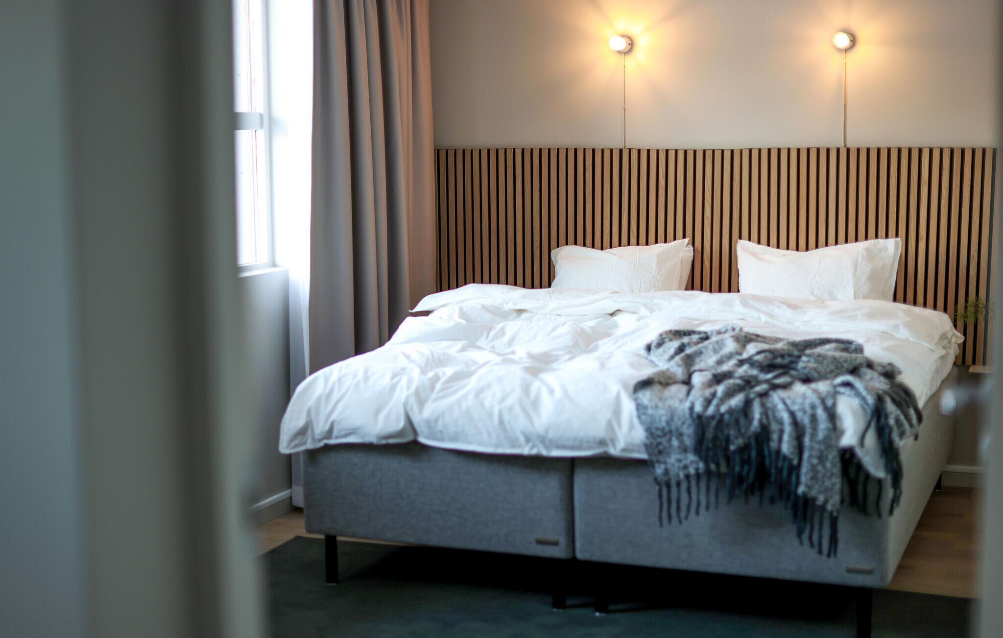Une chambre d’hôtel moderne avec un grand lit, deux tables de chevet, une tête de lit en lattes de bois et deux lampes fixées au mur.