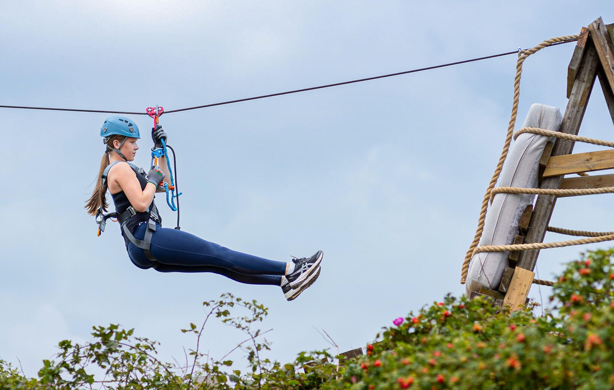 A person zip-lining with safety gear, ropes, and a wooden structure in the background.