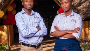 Two individuals standing in front of a rustic building with a thatched roof, wearing uniforms with logos. - The Giraffe Cottages Restaurant and Bar (Nairobi)