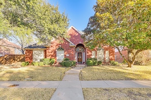 A brick house with a front entrance, a pathway leading to the door, and a small front yard with trees and shrubs.