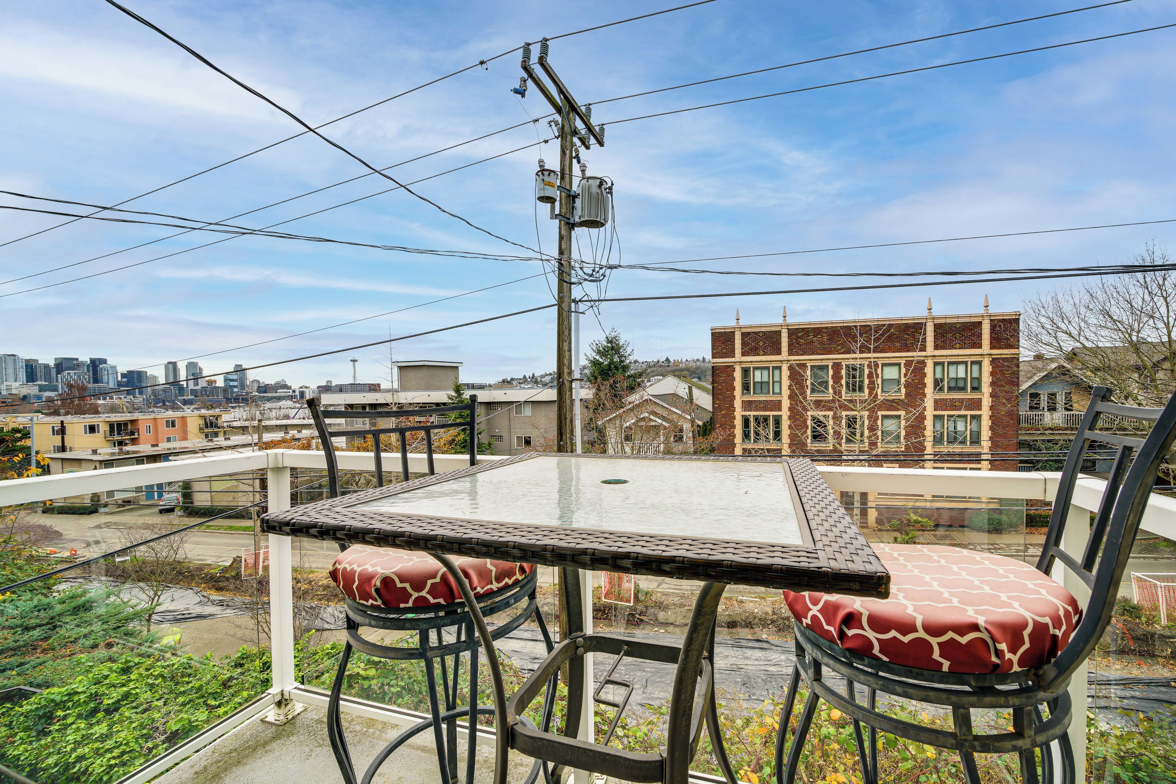A balcony with a glass table and chairs, overlooking a cityscape with buildings and a clear sky.