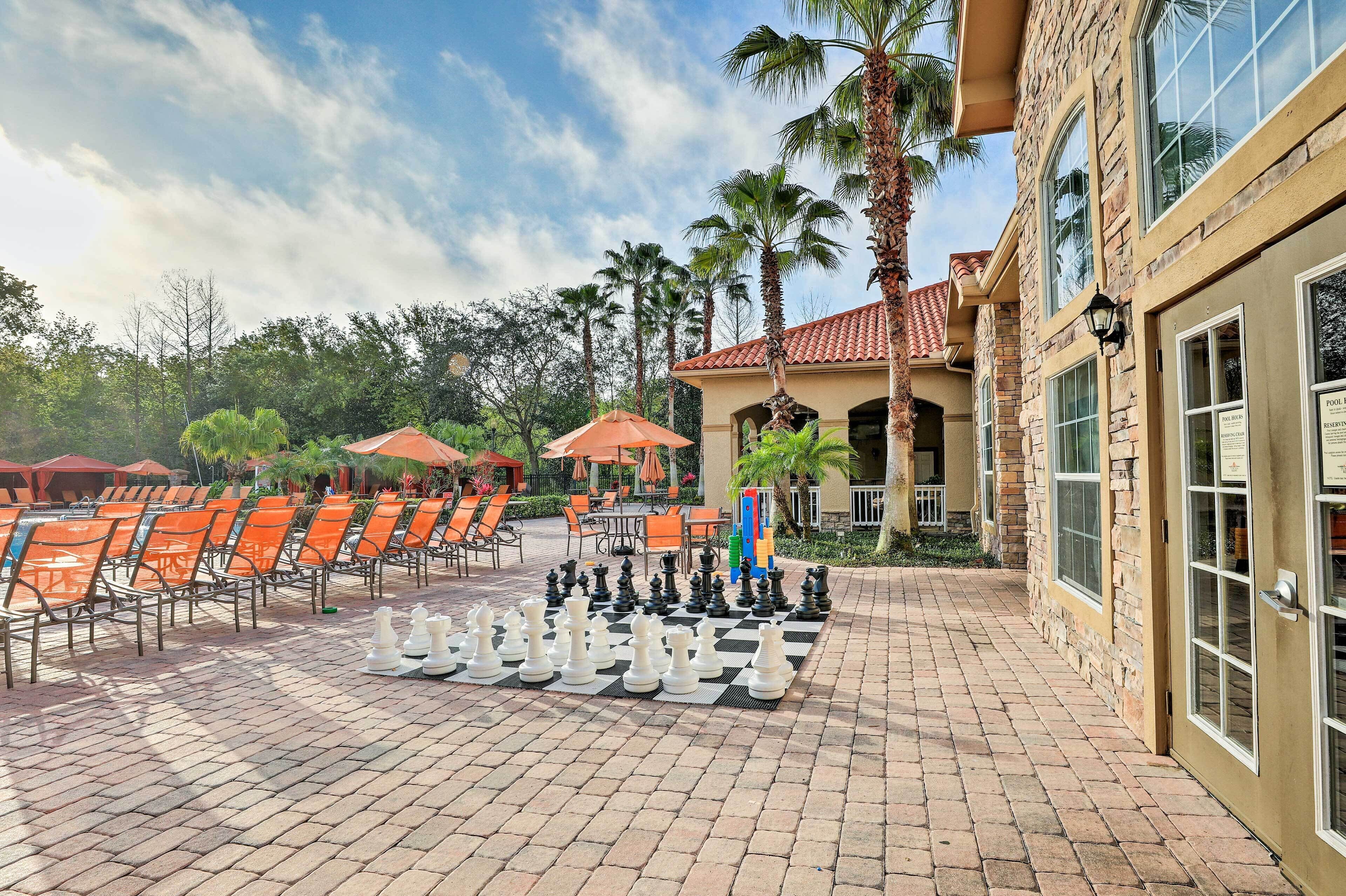 A chessboard set up on a paved patio with orange chairs and umbrellas, palm trees, and a building with a tiled roof.