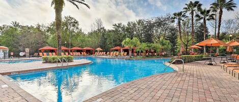 A swimming pool with lounge chairs and umbrellas, surrounded by palm trees and a brick walkway.