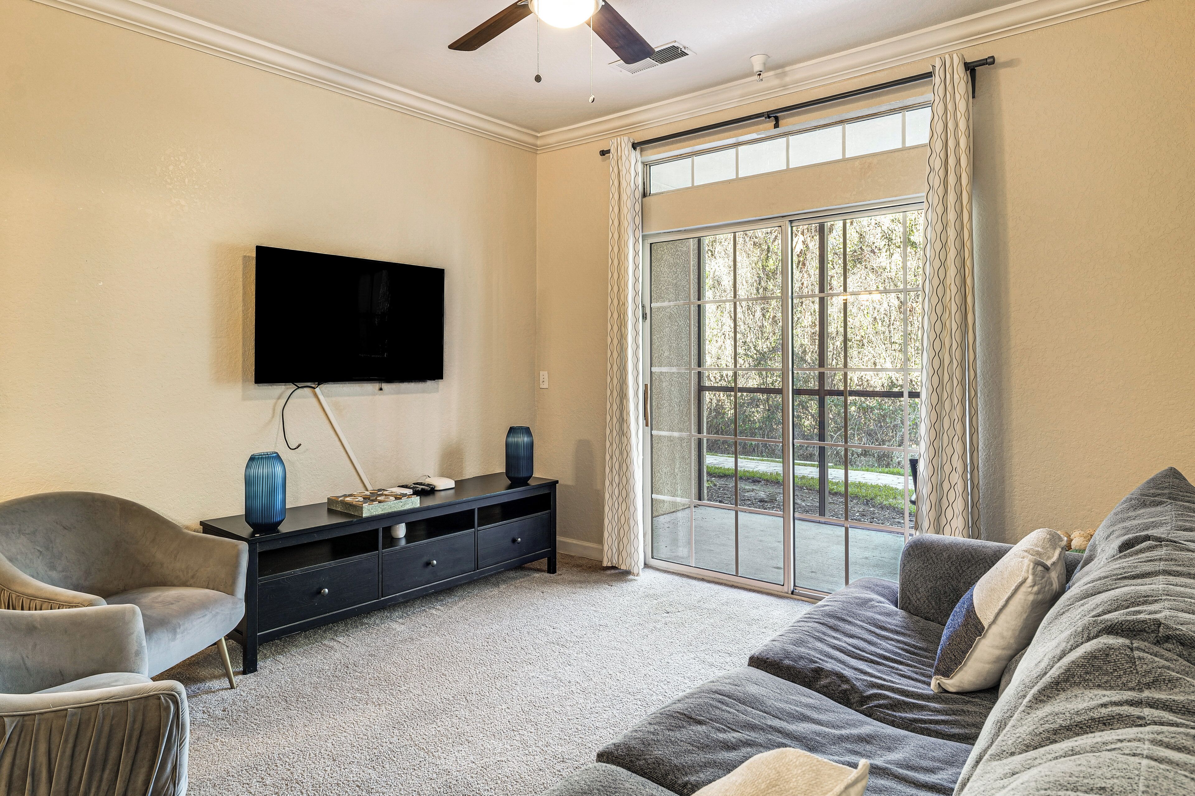 A living room with a flat-screen TV, a sofa, a chair, and a large window with curtains.