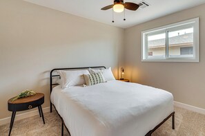 A bedroom with a bed, a round side table with a lamp, a ceiling fan, and a window with a view of buildings.