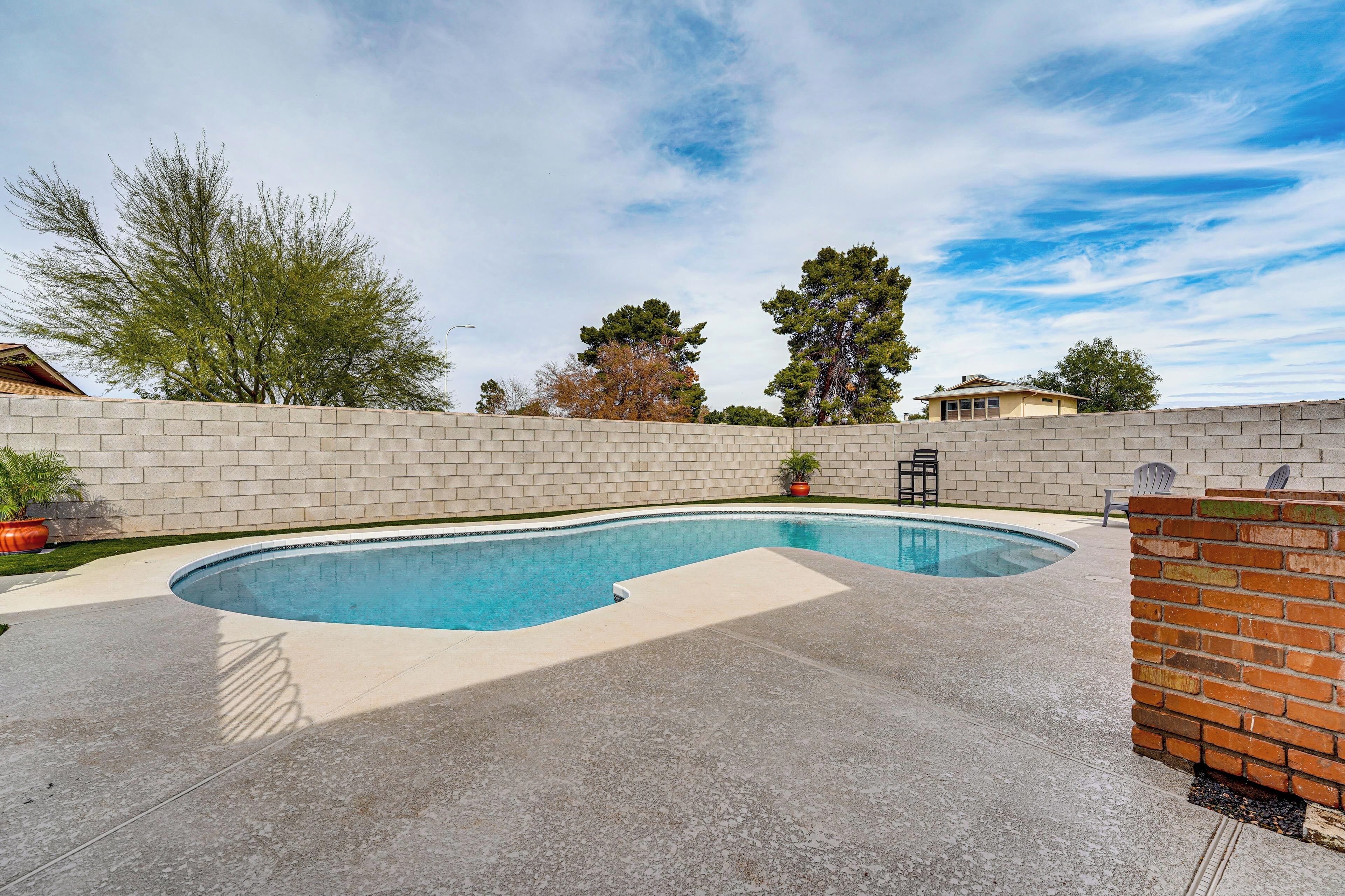 A swimming pool with a brick wall and a concrete deck, surrounded by trees and a clear sky.