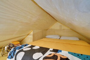 Ein kleines, gemütliches Schlafzimmer im Dachgeschoss mit schrägen Wänden, einem Bett mit bunter Decke sowie einigen Büchern und einem kleinen Tisch mit Lampe.