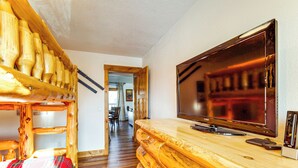 En säng med våningssäng i trä, täckt med ett rutigt överkast, en tv på ett träställ och en hall som leder till ett matområde.
