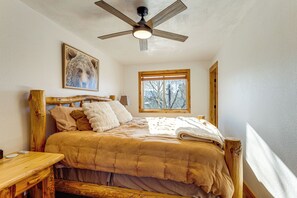 Une chambre avec un lit en bois, un ventilateur de plafond, une fenêtre donnant sur des arbres et un tableau accroché au mur.