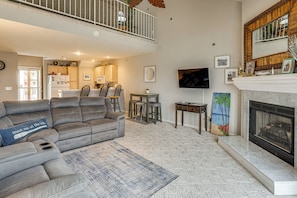 Ein Wohnzimmer mit grauem Ecksofa, einem Kamin, einer Essecke mit Stühlen, einer Küche im Hintergrund und einem Balkon mit Geländer.