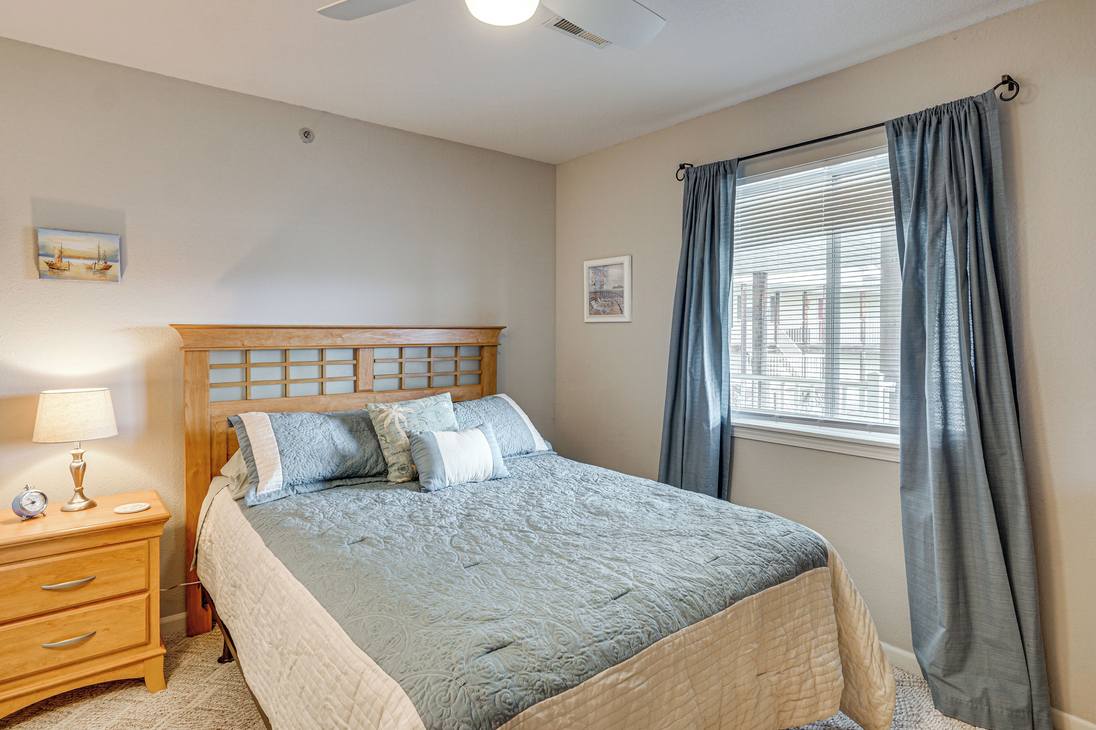 Un dormitorio con cama, mesita de noche, lámpara, ventana con cortinas y ventilador de techo.