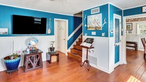 Ein Wohnzimmer mit blauer Wand, Holzboden, einem an der Wand montierten Fernseher, einem Deckenventilator, einer Treppe und einem kleinen Tisch mit einer Pflanze.