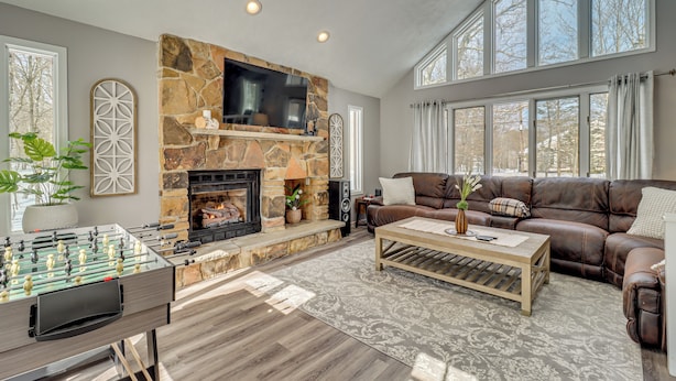 A living room with a stone fireplace, a television mounted above it, a brown leather sofa, and a foosball table. - Contemporary House in Lakeside Community! Poconos (Albrightsville)
