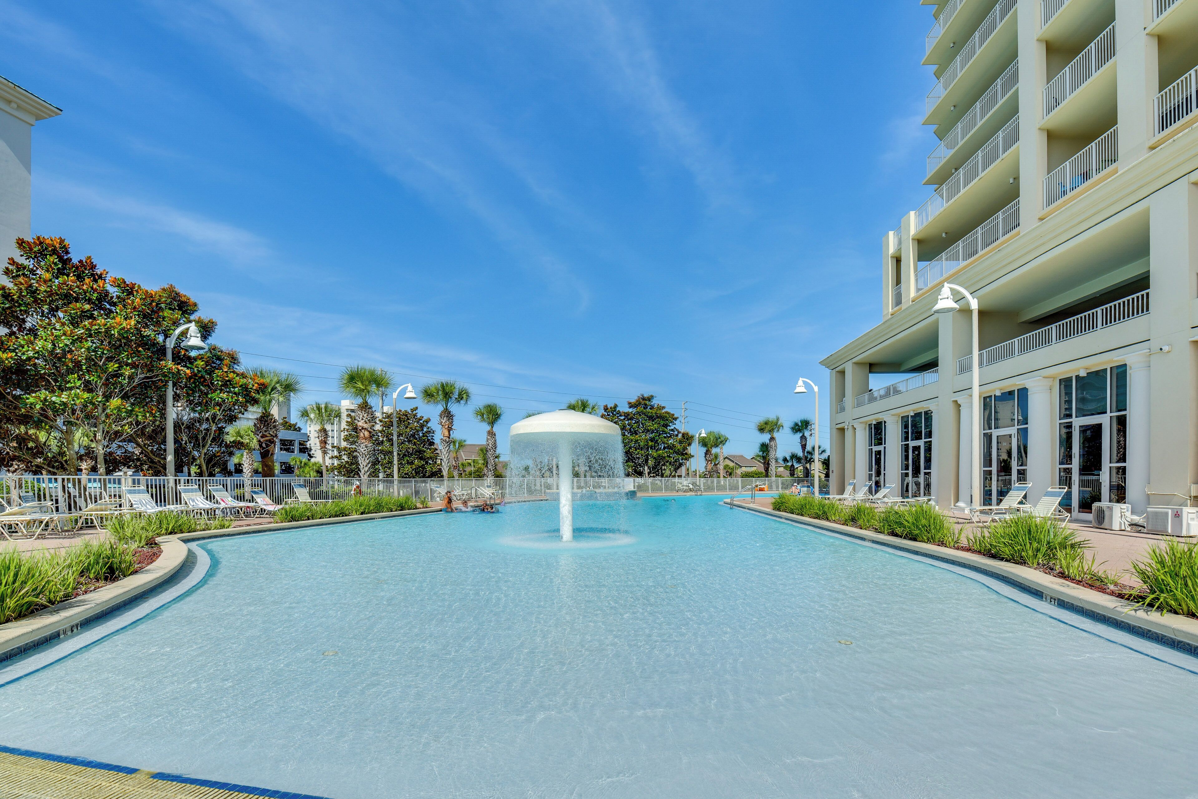 A swimming pool with a central fountain, surrounded by lounge chairs and palm trees, with a hotel building in the background.