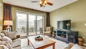 A living room with a sofa set, a wooden coffee table, a television, and a large window with a balcony. - Steps to Sea! Snowbird Haven in Miramar Beach (Miramar Beach)