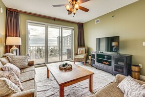A living room with a sofa set, a wooden coffee table, a television, and a large window with a balcony. - Steps to Sea! Snowbird Haven in Miramar Beach (Miramar Beach)