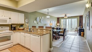 A spacious kitchen with white cabinets, a granite countertop, and a dishwasher. There is a dining area with a table and chairs, and a view of a balcony with a railing. - Steps to Sea! Snowbird Haven in Miramar Beach (Miramar Beach)