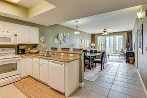 A spacious kitchen with white cabinets, a granite countertop, and a dishwasher. There is a dining area with a table and chairs, and a view of a balcony with a railing. - Steps to Sea! Snowbird Haven in Miramar Beach (Miramar Beach)