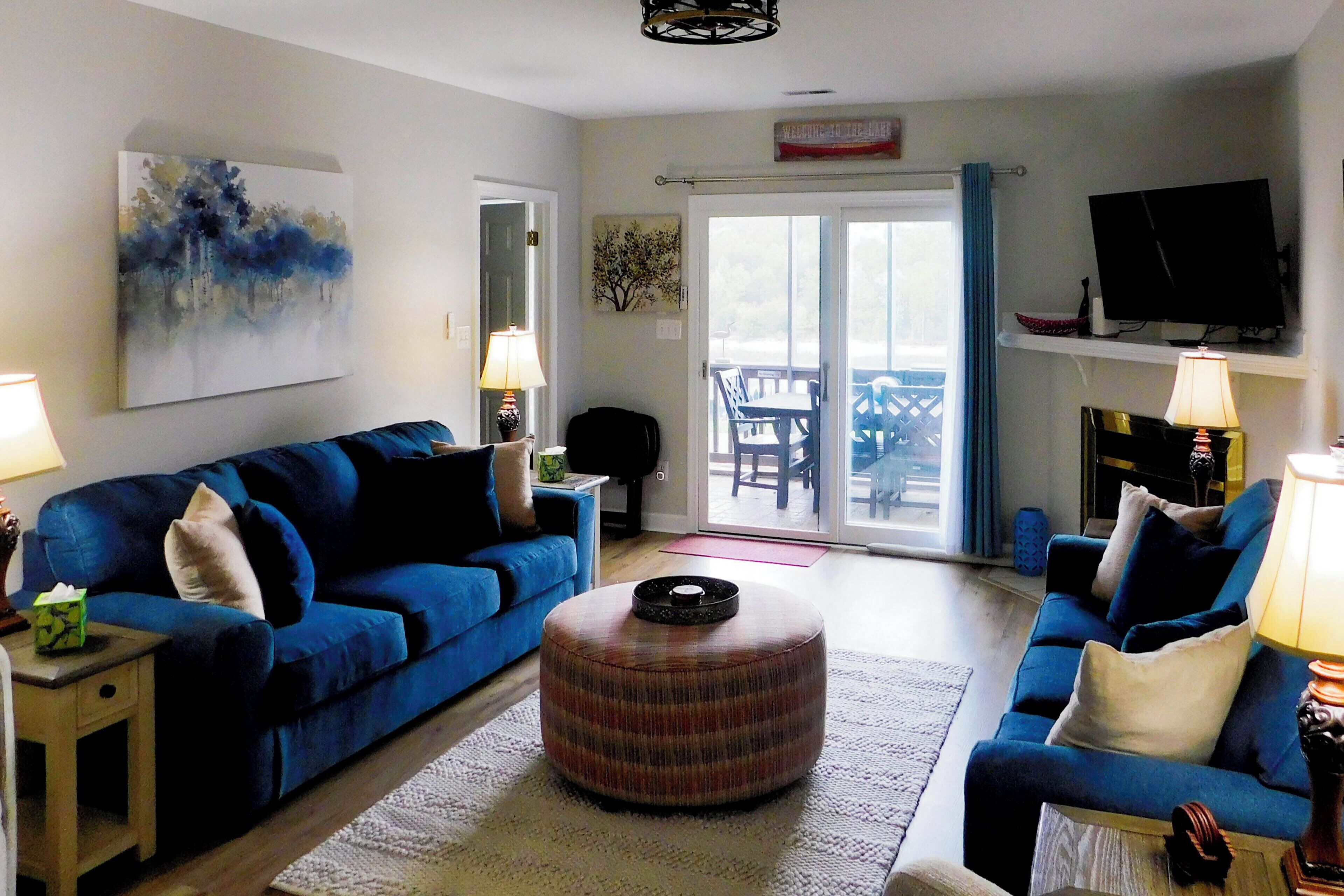 Ein Wohnzimmer mit einem blauen Sofa, einem runden Hocker, einem Fernseher und einem Bild an der Wand.