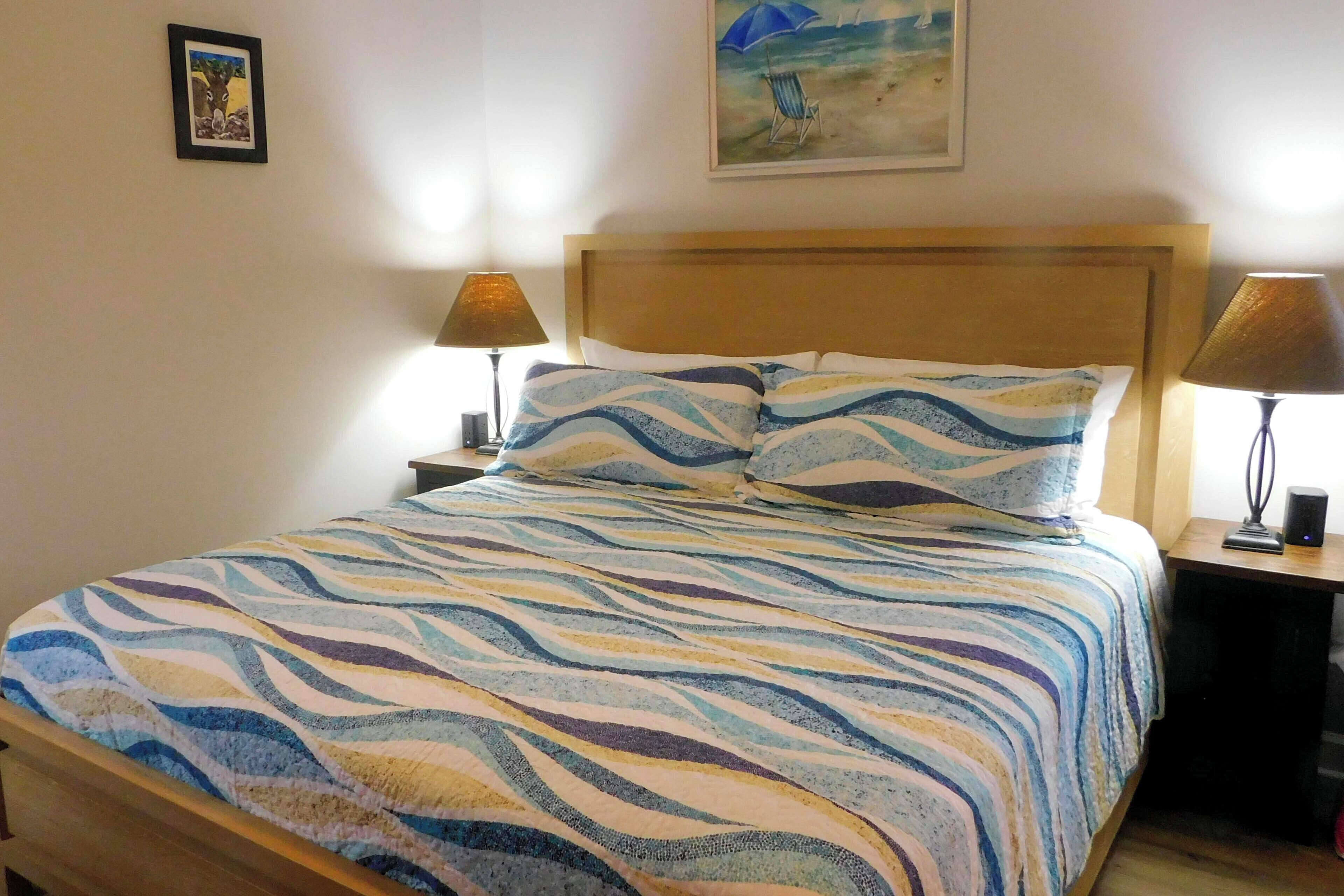 Ein Bett mit gemustertem Überwurf, zwei Nachttische mit Lampen, ein gerahmtes Bild an der Wand und ein Gemälde über dem Bett.
