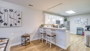 A modern kitchen with white cabinetry, a central island with bar stools, pendant lighting, and a large wall clock. - Dog-friendly Condo on Smith Mountain Lake (Huddleston)