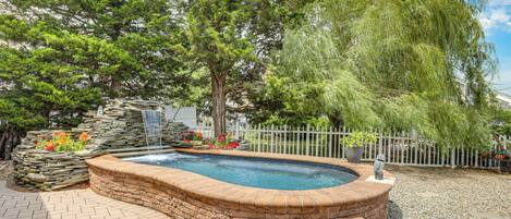 A swimming pool with a stone edge, surrounded by a brick patio, a white fence, and trees.