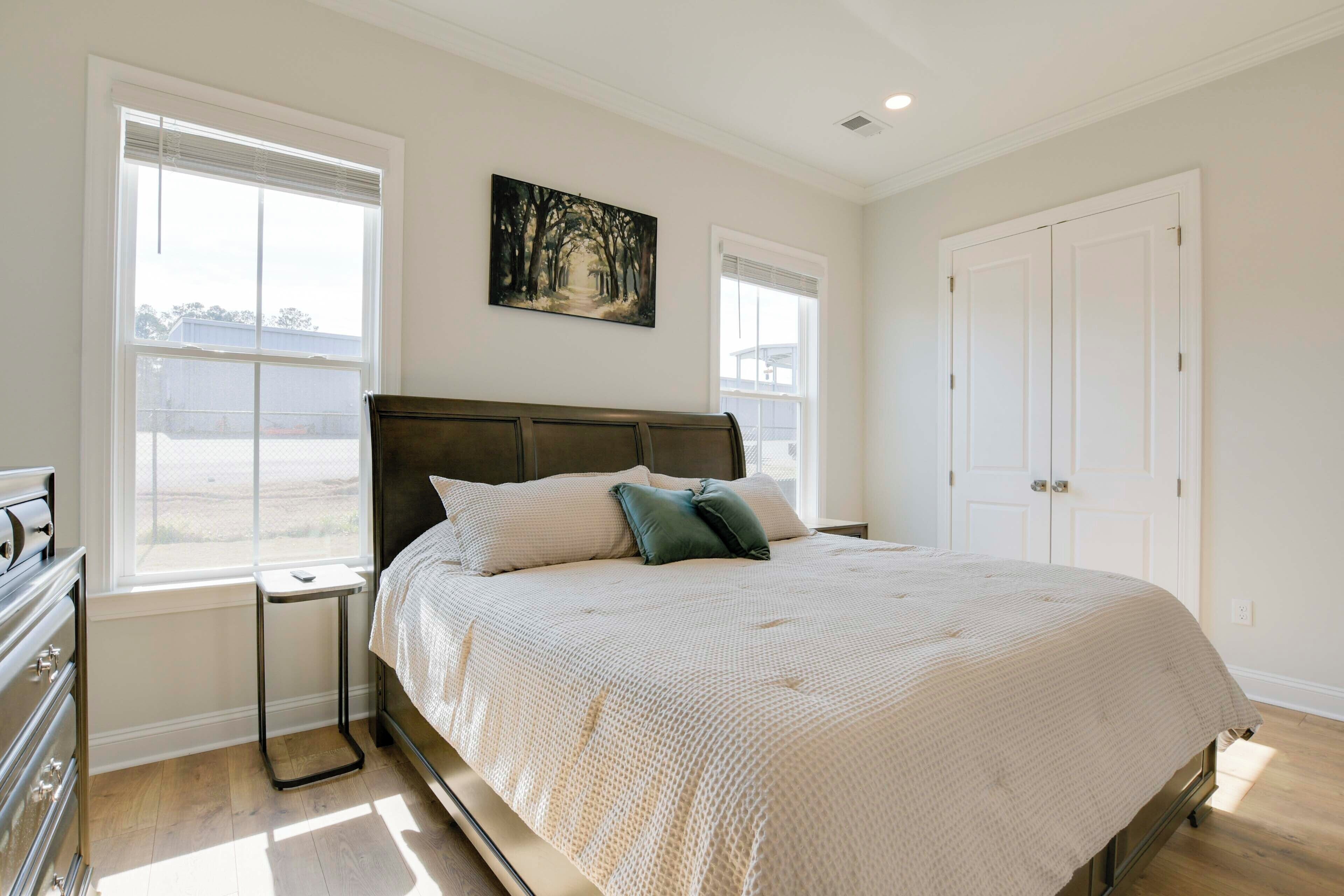 A bedroom with a large bed, two windows, a nightstand, a dresser, and a painting on the wall.