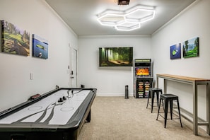 A room with a pool table, a foosball table, and a video game machine. There are two paintings on the wall and a large forest painting above the foosball table.