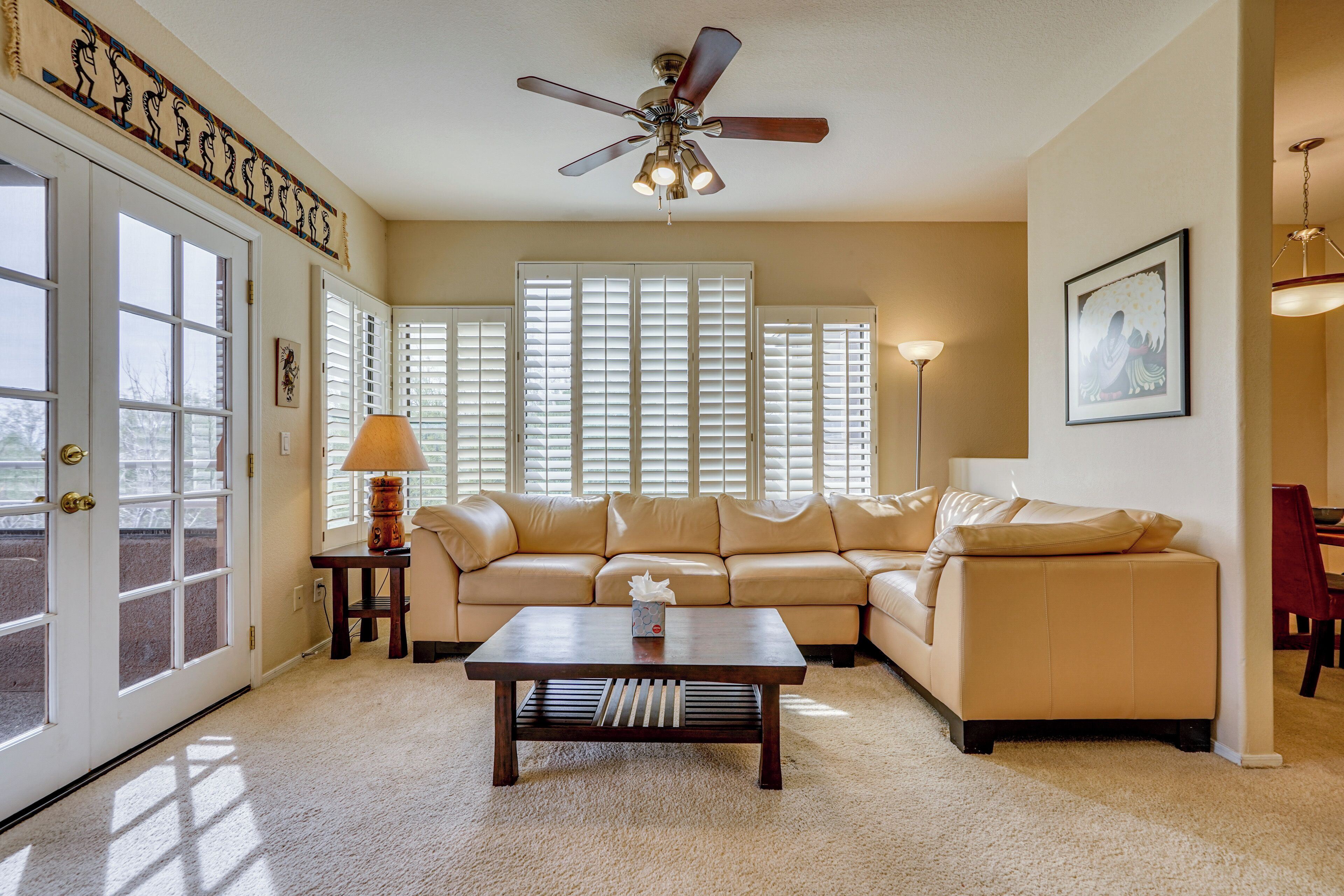 A living room with a beige sofa, a wooden coffee table, a ceiling fan, and a large window with blinds.
