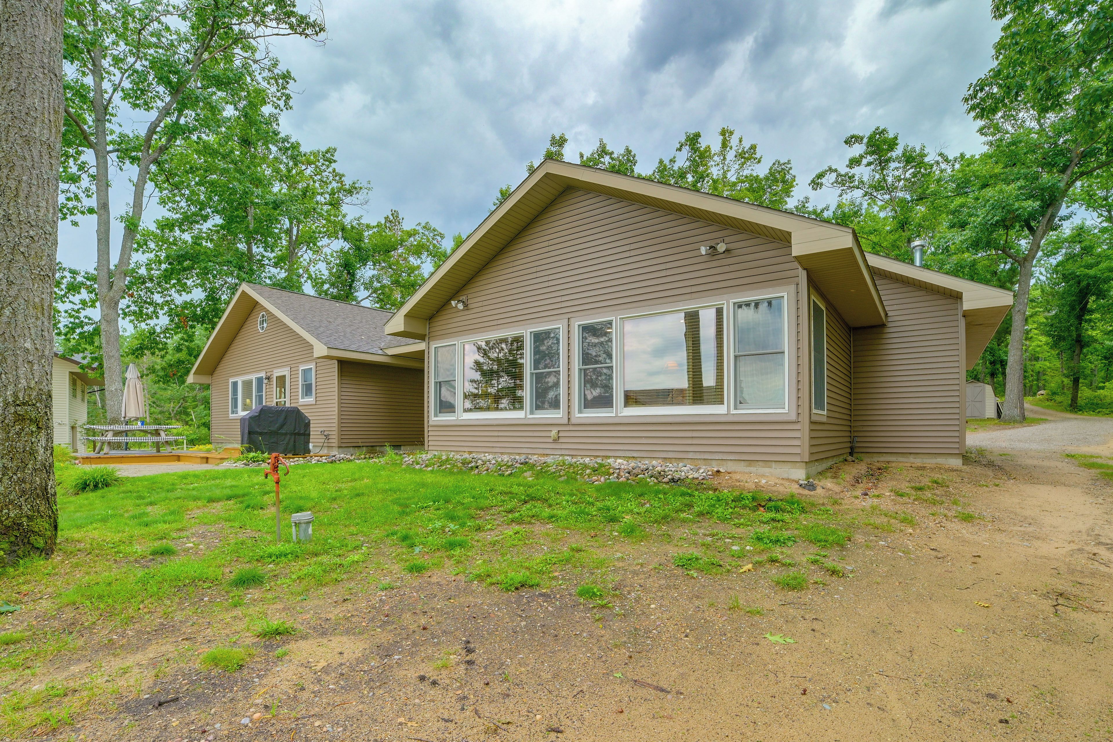 A two-story cabin with a large window on the front, surrounded by trees and a gravel driveway.