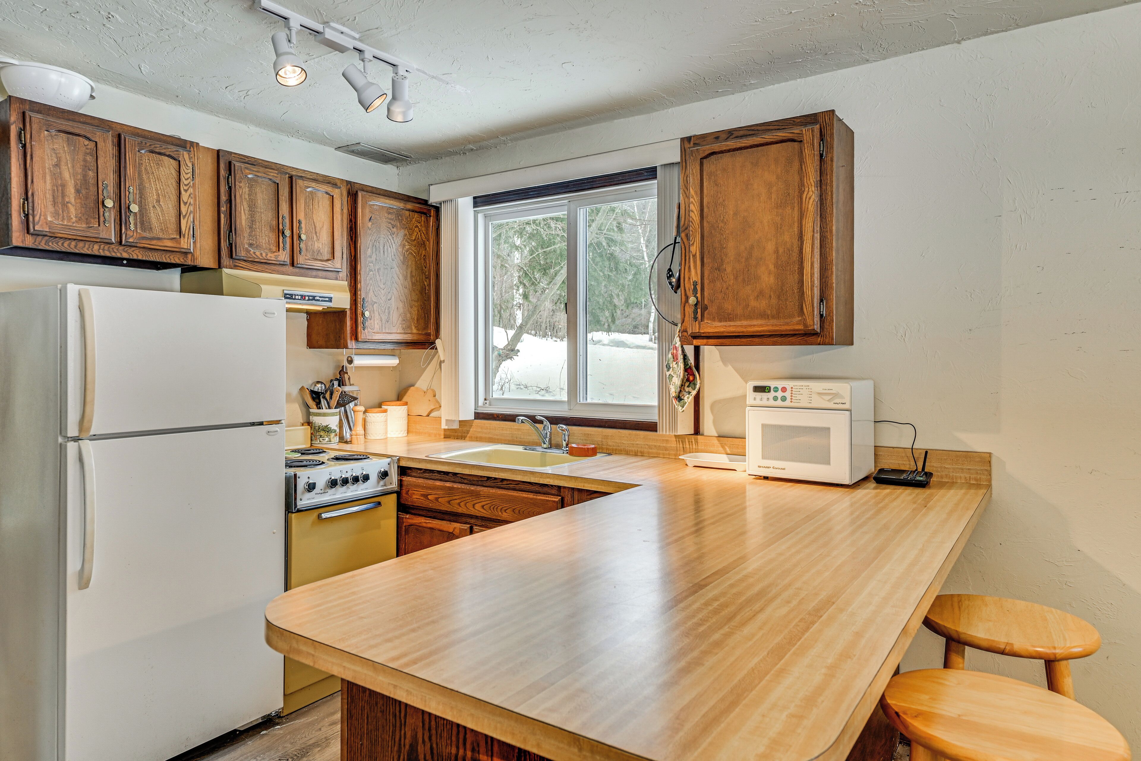 Une cuisine avec des meubles en bois, un réfrigérateur blanc, une cuisinière, un four à micro-ondes et une fenêtre donnant sur des arbres.