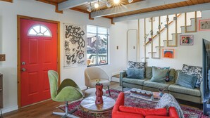 Una sala de estar con una puerta roja, una silla verde, un sillón color beige y un sofá azul, además de una alfombra estampada.