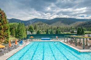 Una piscina con sillones de descanso y sombrillas, rodeada por una valla y árboles, con montañas al fondo.