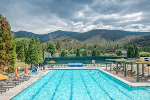 Una piscina con sillones de descanso y sombrillas, rodeada por una valla y árboles, con montañas al fondo.