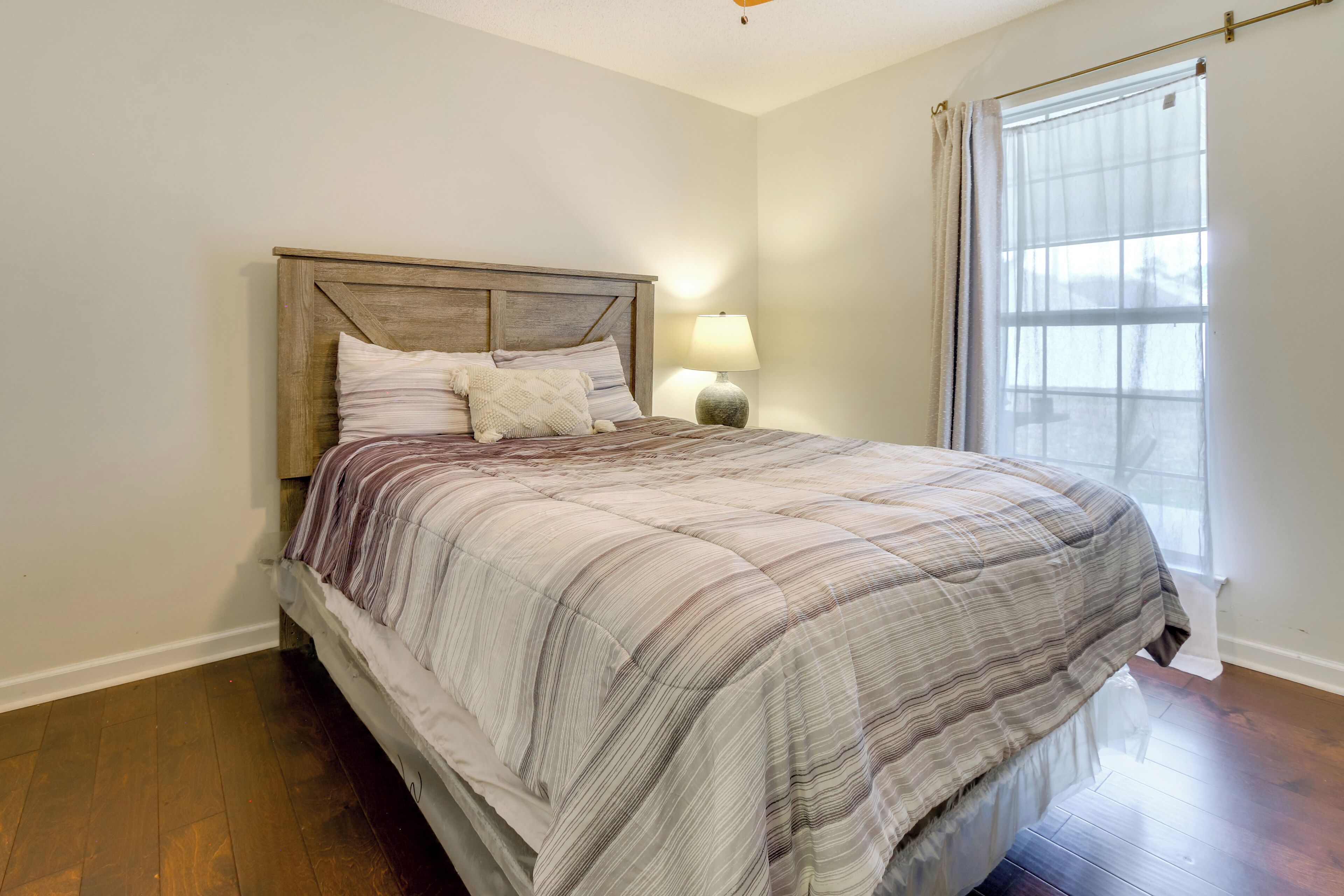 A bedroom with a wooden headboard, a bed with a striped blanket, a bedside lamp, and a window with a sheer curtain.
