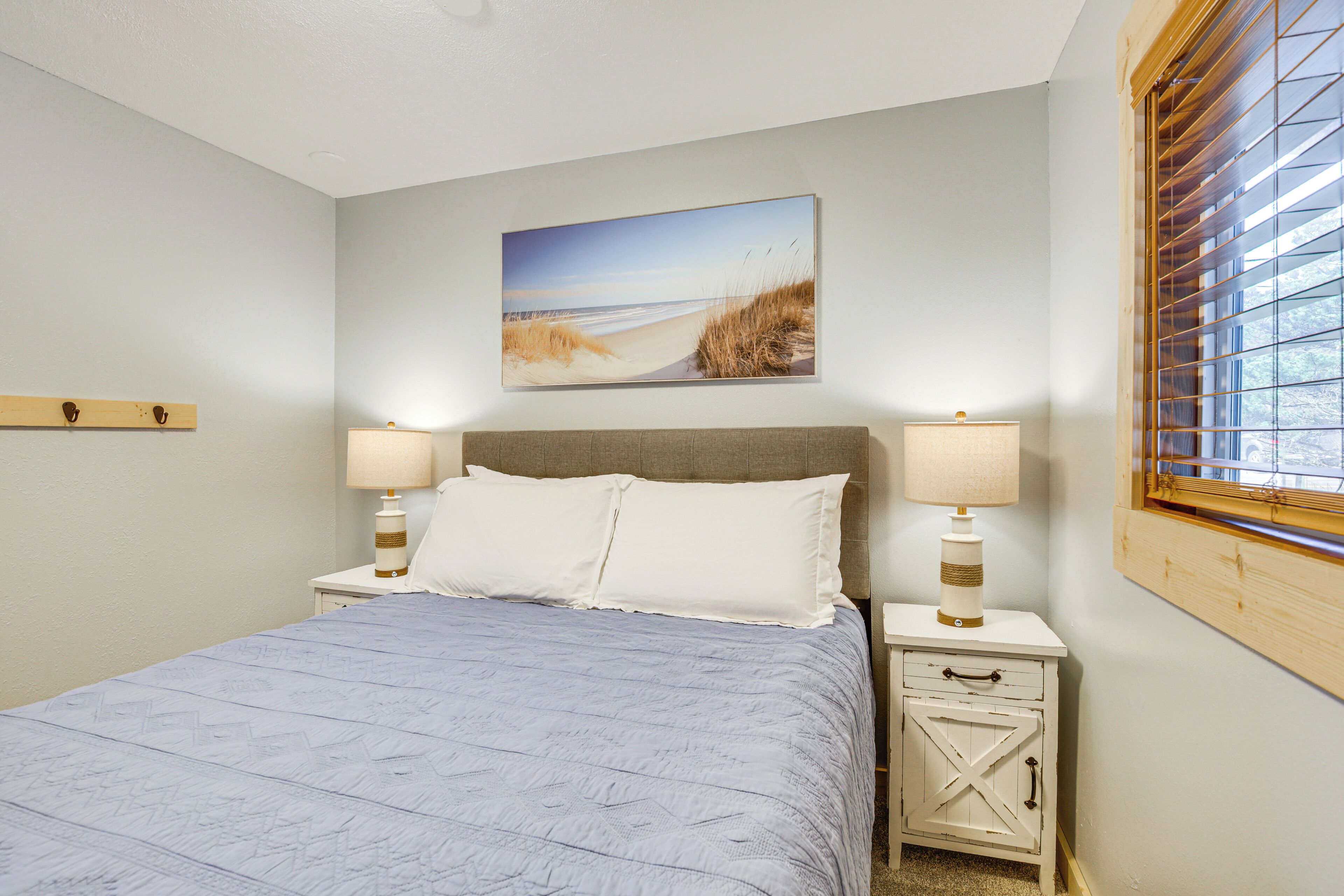 A bedroom with a bed, two bedside tables with lamps, a nightstand, a wooden headboard, a window with blinds, and a beach-themed painting on the wall.