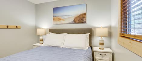 A bedroom with a bed, two bedside tables with lamps, a nightstand, a wooden headboard, a window with blinds, and a beach-themed painting on the wall.