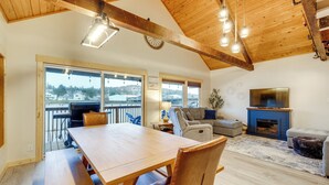 Un vaste espace de vie doté d’un plafond en bois, d’une table à manger avec des bancs, d’une cheminée et de grandes fenêtres offrant une vue sur l’extérieur.