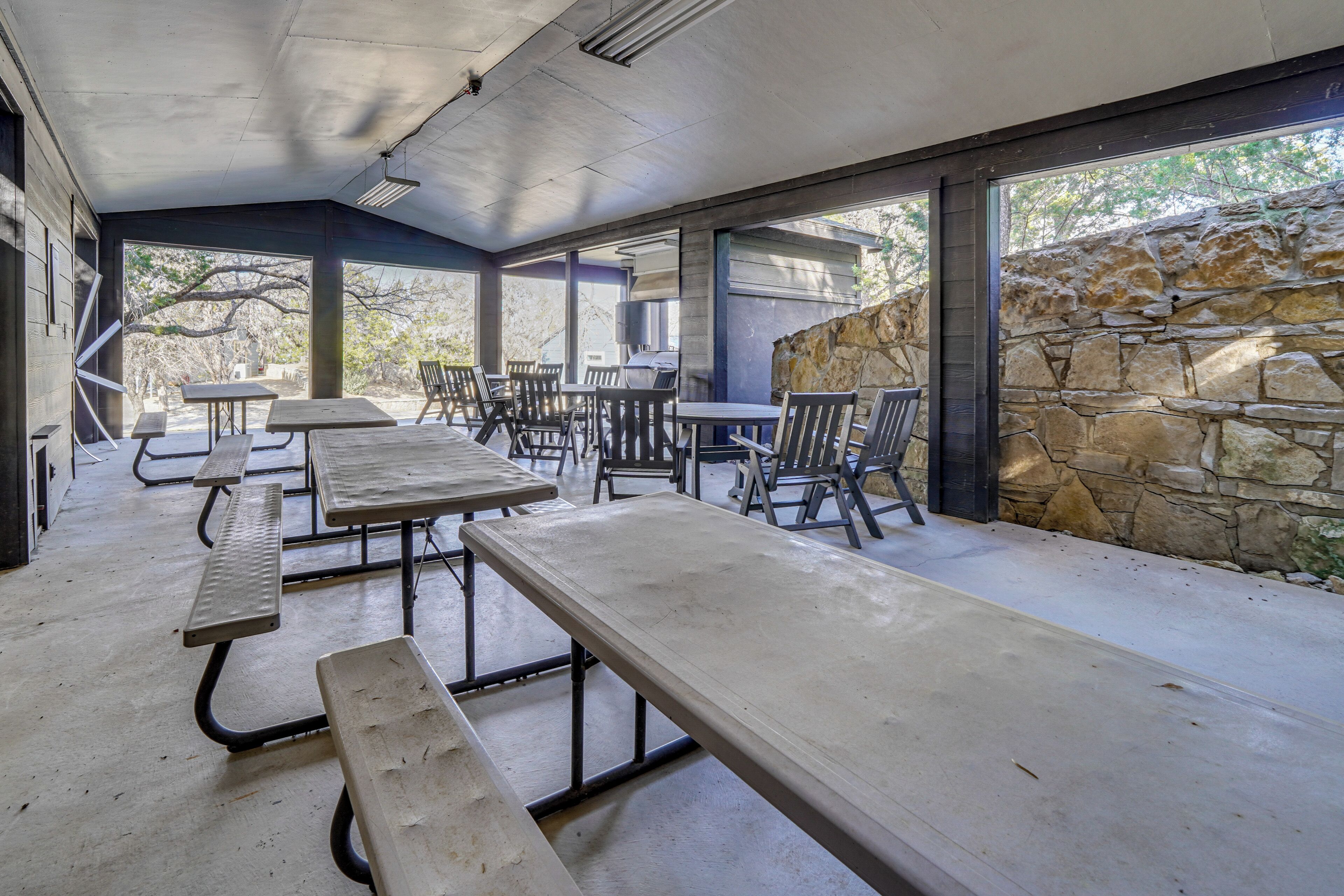 An outdoor patio area with picnic tables and chairs, stone walls, and large windows.