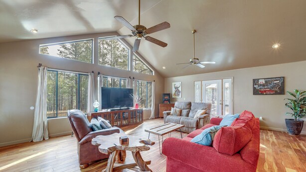 A spacious living room with a red sofa, a brown leather chair, and a wooden coffee table. There are two ceiling fans, large windows, and a television on a stand. - Great for Families! Woodsy Haven in Happy Jack (Happy Jack)
